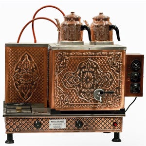 Może przedstawiać: Miedziany ekspres do herbaty z dwoma dekoracyjnymi czajniczkami. Ekspres do herbaty ma ozdobny, wytłoczony wzór i mały palnik. Panel sterowania znajduje się po prawej stronie. Ekspres do herbaty stoi na białej powierzchni.