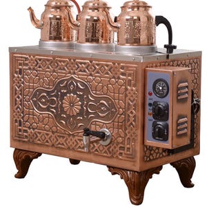 Op de afbeelding: Een koperkleurige theemaker met drie decoratieve theepotten. De theemaker heeft een sierlijke, patroonfront en staat op vier gesneden houten poten. Het heeft een bedieningspaneel en een kraan.