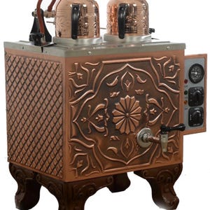 Puede incluir: Una tetera turca de color cobre con dos teteras decorativas. La tetera tiene patrones florales ornamentados, un grifo y un panel de control. La base tiene patas decorativas. La tetera está diseñada para hacer té.