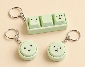 May include: Three light green keychains featuring minimalist facial expressions. One keychain is a rectangular base with three square keys, while the other two are circular buttons. Each piece has a metal ring attached by a silver chain.