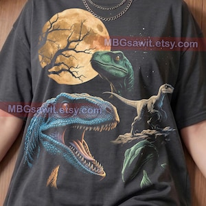 May include: A charcoal grey t-shirt featuring a graphic print of three dinosaurs, a large yellow moon, and the text MBGsawit.etsy.com. The design includes a blue raptor with an open mouth, a green raptor head, and a smaller raptor standing on a rock.