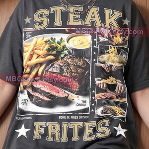 May include: A charcoal gray t-shirt featuring a large graphic print of a steak and fries meal. The text reads STEAK, FRITES, JUICY CUTS, THE CHOICE, FLAVOR KING, BONE IN, FRIES ON SIDE, and Pepper Comfort Colors. Smaller images show cooking processes.