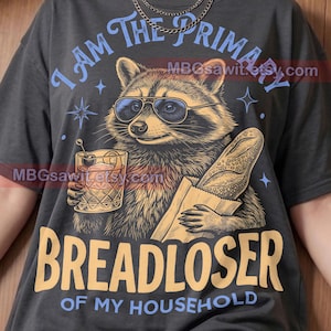May include: A charcoal gray t-shirt featuring a graphic of a raccoon wearing sunglasses, holding a cocktail and a baguette. The text reads I AM THE PRIMARY BREADLOSER OF MY HOUSEHOLD. The shirt includes the Pepper Comfort Colors brand logo.