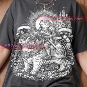 May include: Dark gray t-shirt featuring a detailed illustration of a frog in knight's armor riding a cat. The design includes a castle, floral elements, and the text "Pepper Comfort Colors".