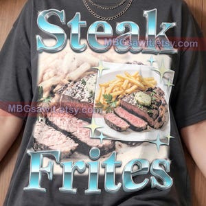 May include: A dark gray t-shirt featuring a graphic print of a sliced steak and french fries. The word "Steak" appears in large, metallic blue letters at the top, and "Frites" in the same style at the bottom. The text "Pepper Comfort Colors" is visible.