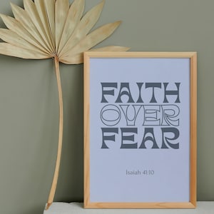 Puede incluir: Una impresión enmarcada con las palabras "FAITH OVER FEAR" en letras grises. La impresión está en un marco de madera clara y descansa sobre una superficie blanca. A la izquierda del marco hay un abanico de hojas de palma secas.