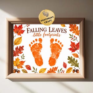 May include: A framed print featuring the text FALLING LEAVES little footprints in brown script. Two orange baby footprints are centered on a white background, surrounded by a border of watercolor autumn leaves, berries, and acorns.