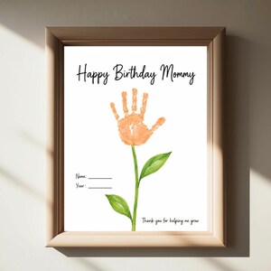 Op de afbeelding: Een ingelijste print met de tekst Happy Birthday Mommy bovenaan en Thank you for helping me grow onderaan. Een centrale oranje handafdruk vormt een bloem op een groene steel met twee bladeren. Bevat lijnen voor Name en Year.