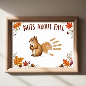 以下が含まれることがあります： どんぐりを持つ茶色のリス、ベージュの手形、そして「NUTS ABOUT FALL」という文字が描かれた額入りプリント。四隅には色鮮やかな秋の葉、ベリー、どんぐりが装飾されており、全体が白い背景に配置されています。