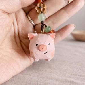 Peut inclure: Un porte-clés en forme de cochon rose sculpté à la main, avec un visage souriant, de petites oreilles et un museau rond. Le porte-clés est orné de perles, d'une fleur en bois et de pierres vertes.