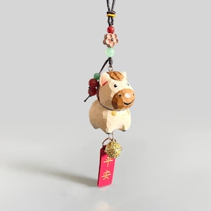May include: A carved wooden horse charm with a brown and cream color scheme, accented with a red tag and a gold bell. The charm is suspended from a black cord with decorative beads and a flower.