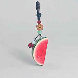 May include: A watermelon slice charm with a beaded cord. The charm features a red interior with black seeds, green rind, and a decorative beaded strand with a flower and other beads. The charm is suspended from a black cord.