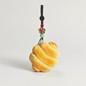 May include: A carved wooden ornament in the shape of a swirl, painted in shades of yellow and orange. It hangs from a black cord with decorative beads and a flower charm. The background is plain white.
