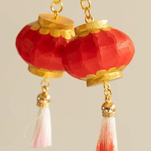 Puede incluir: Un par de pendientes de linterna rojos y dorados con cadenas doradas y borlas blancas y rojas. Los pendientes tienen un aspecto de madera tallada y están diseñados al estilo asiático tradicional.