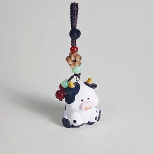 May include: A white and black cow figurine with a pink nose and yellow horns, attached to a beaded string with a flower and berry accents. The cow has black spots and is sitting down.