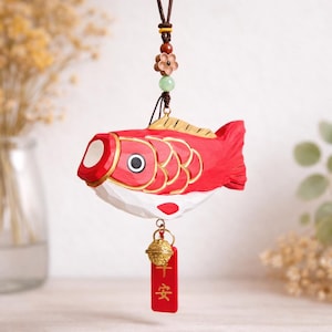 May include: A red and white carved wooden fish ornament with gold accents, a small bell, and a red tag with Chinese characters. The fish is suspended by a brown cord with decorative beads and a flower.