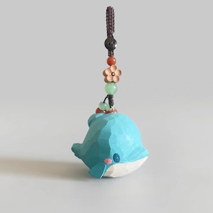 May include: A carved wooden dolphin keychain in light blue with a white belly and pink accents. The dolphin is suspended from a brown braided cord with decorative beads, including a wooden flower and green and red stones. The keychain is a charming accessory.