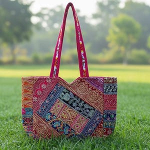 May include: A colorful tote bag with a patchwork design. The bag features a variety of patterns and colors, including red, blue, gold, and green. The handles are a solid magenta color. The bag is sitting on a bed of green grass.