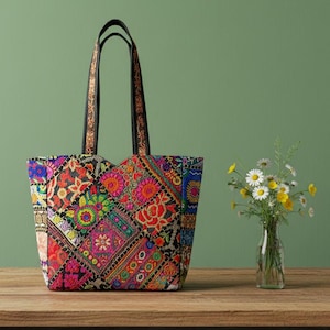May include: A colorful tote bag with a patchwork design. The bag features a variety of embroidered patterns in red, orange, blue, and green. It has black handles with decorative stitching and is set on a wooden surface.