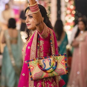 Puede incluir: Un bolso bordado en rosa y dorado con un asa de arcoíris y un diseño de pavo real. El bolso lo lleva una persona que viste un vestido y brazaletes bordados en rosa y dorado. El diseño del bolso incluye bordados coloridos y espejos.