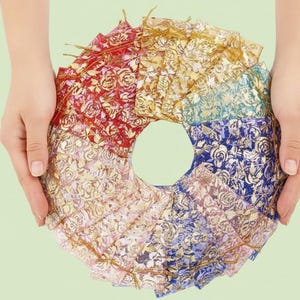 Puede incluir: Una disposición circular de pequeñas bolsas de tela coloridas con patrones florales dorados. Las bolsas son en tonos de rojo, dorado, azul y rosa, y están sostenidas por dos manos sobre un fondo verde claro. Cada bolsa tiene un cordón dorado.