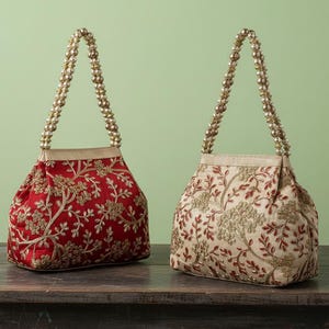 Op de afbeelding: Twee sierlijke handtassen met kralen handvatten. Eén is rood met gouden bloemenborduurwerk, de andere is beige met vergelijkbaar borduurwerk. Beide tassen hebben gouden kralen handvatten en staan op een donkerhouten oppervlak.