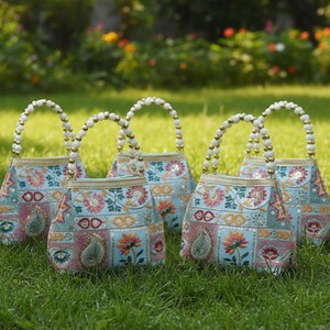Könnte beinhalten: Fünf bestickte Handtaschen mit Perlenhenkeln auf Gras. Die Taschen haben einen hellblauen Hintergrund mit Blumen- und Paisley-Mustern in Rosa, Orange und Grün. Goldbesatz akzentuiert die Oberseite jeder Tasche.