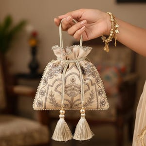 May include: A beige, embroidered drawstring bag with a decorative floral pattern. The bag features a beaded trim, a drawstring closure, and two tassels. The bag is held by a person.
