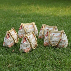 May include: Five small, cream-colored fabric handbags with floral patterns in red and peach. Each bag has a gold beaded handle and gold trim. The bags are arranged on a bed of green grass.