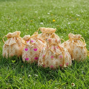 Può includere: Una collezione di sacchetti con coulisse color crema con ricami floreali dorati e rosa. I sacchetti sono disposti su erba verde, con chiusura a coulisse con perline e finiture decorative.