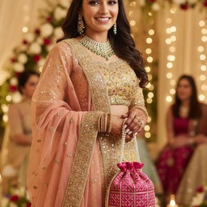 Op de afbeelding: Een vrouw in een traditionele Indiase trouwoutfit. Ze draagt een goudkleurige en perzikgekleurde geborduurde top en rok, met een bijpassende perzikkleurige sjaal. Ze houdt een roze trekkoordtas met een geometrisch patroon vast. Ze draagt ook een ketting en oorbellen.
