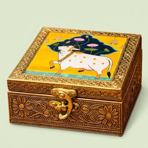 May include: A square, gold-colored decorative box with a hinged lid and clasp. The lid features a painting of a white cow with orange spots against a yellow background. The box is adorned with floral and geometric patterns.