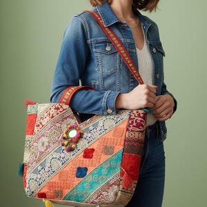 Puede incluir: Un bolso tote colorido con un diseño de patchwork. El bolso presenta una variedad de patrones y colores, incluyendo rojo, naranja, turquesa y dorado. Tiene un espejo decorativo y borlas. La correa es roja con un diseño estampado.
