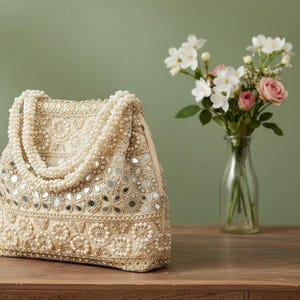 May include: A beige beaded handbag with a pearl handle and mirrored details. The bag is adorned with small pearls and floral patterns. A bouquet of white and pink flowers is in a clear vase in the background.