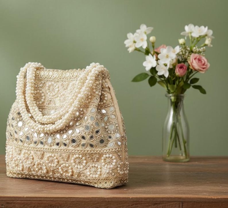 May include: A beige beaded handbag with a pearl handle and mirrored details. The bag is adorned with small pearls and floral patterns. A bouquet of white and pink flowers is in a clear vase in the background.
