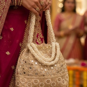 May include: A gold-toned, embellished handbag with a pearl handle. The bag has detailed embroidery and small mirror accents. The handle is made of multiple strands of white pearls. The background shows a person wearing a red garment with gold embroidery.