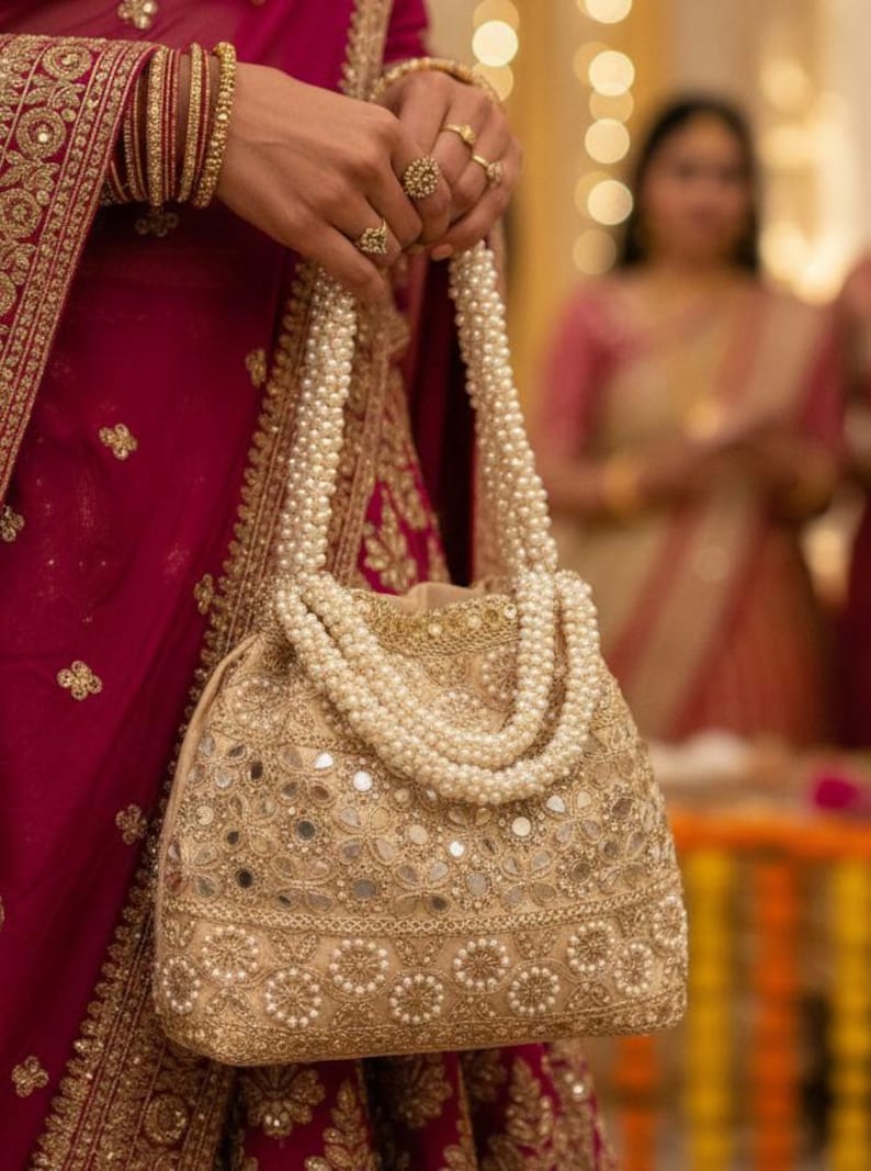 May include: A gold-toned, embellished handbag with a pearl handle. The bag has detailed embroidery and small mirror accents. The handle is made of multiple strands of white pearls. The background shows a person wearing a red garment with gold embroidery.