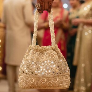 May include: A gold-coloured, beaded drawstring bag with a pearl handle. The bag features small, reflective mirror accents and intricate floral patterns. The handle is made of white pearls.