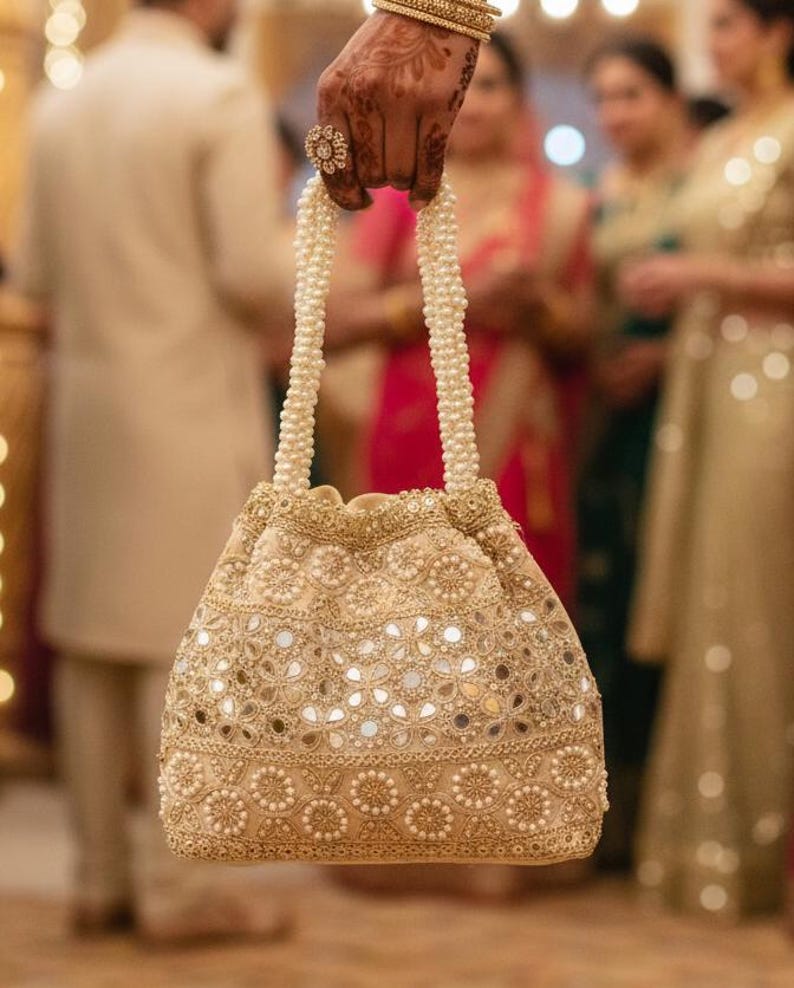 May include: A gold-coloured, beaded drawstring bag with a pearl handle. The bag features small, reflective mirror accents and intricate floral patterns. The handle is made of white pearls.