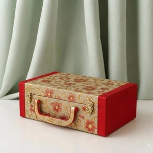 Könnte beinhalten: Eine rechteckige Aufbewahrungsbox mit goldfarbenem Deckel und Vorderseite, die mit einem sich wiederholenden Muster aus roten Blumen und goldenen Ranken verziert ist. Die Box hat einfarbig rote Seitenteile, einen passenden roten Griff und zwei goldene Metallverschlüsse.