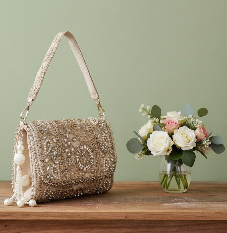 May include: A beige beaded handbag with a long strap and a decorative tassel. The bag is embellished with intricate beadwork and a flap closure. A small bouquet of white and pink roses sits nearby.