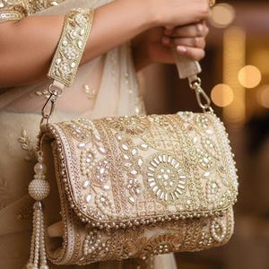 May include: A cream-coloured clutch bag with gold and pearl detailing. The rectangular bag has detailed embroidery, mirrored elements, and a beaded tassel. A matching strap is attached with gold-toned hardware. The bag is held by a person.