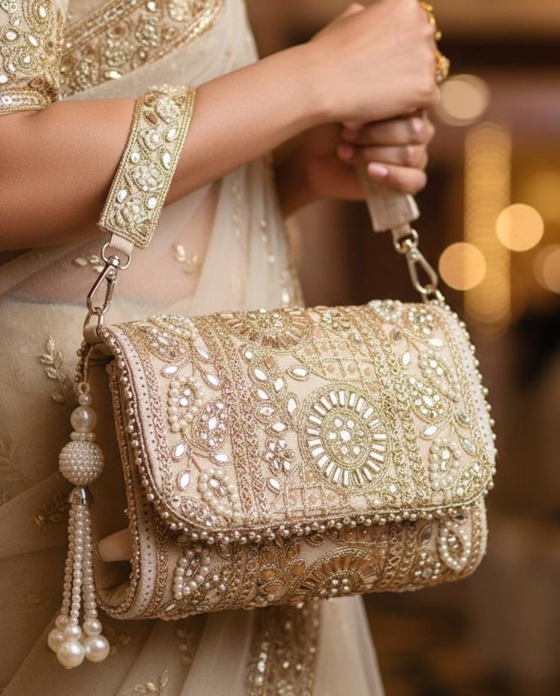 May include: A cream-coloured clutch bag with gold and pearl detailing. The rectangular bag has detailed embroidery, mirrored elements, and a beaded tassel. A matching strap is attached with gold-toned hardware. The bag is held by a person.