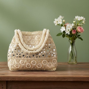 May include: A beige handbag with detailed embroidery, small mirrors, and pearl embellishments. The bag has a beaded handle and sits on a wooden surface. A vase of flowers is in the background.