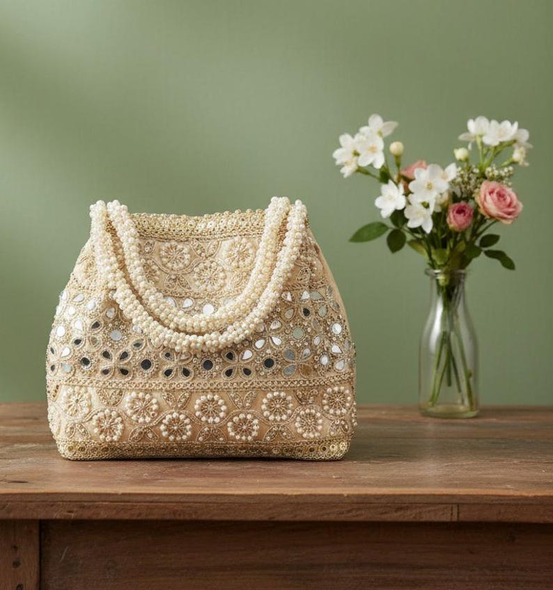 May include: A beige handbag with detailed embroidery, small mirrors, and pearl embellishments. The bag has a beaded handle and sits on a wooden surface. A vase of flowers is in the background.