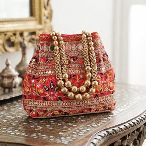 May include: A red bucket bag featuring intricate multi-colored floral embroidery, gold-tone sequin accents, and a decorative handle composed of two strands of large, polished gold-colored beads that drape over the front of the bag.