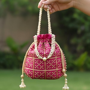 Può includere: Una borsa a coulisse in raso rosa con ricami dorati a forma di griglia. La borsa ha un manico di perle e una grande perla al centro. Nappe e finiture dorate completano il design. La borsa è tenuta in mano.