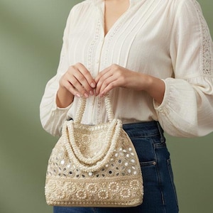 May include: A cream-coloured handbag with intricate beadwork and mirror accents. The bag features a pearl-beaded handle. The bag's design includes floral patterns and a structured shape. The person is wearing a white blouse and blue jeans.