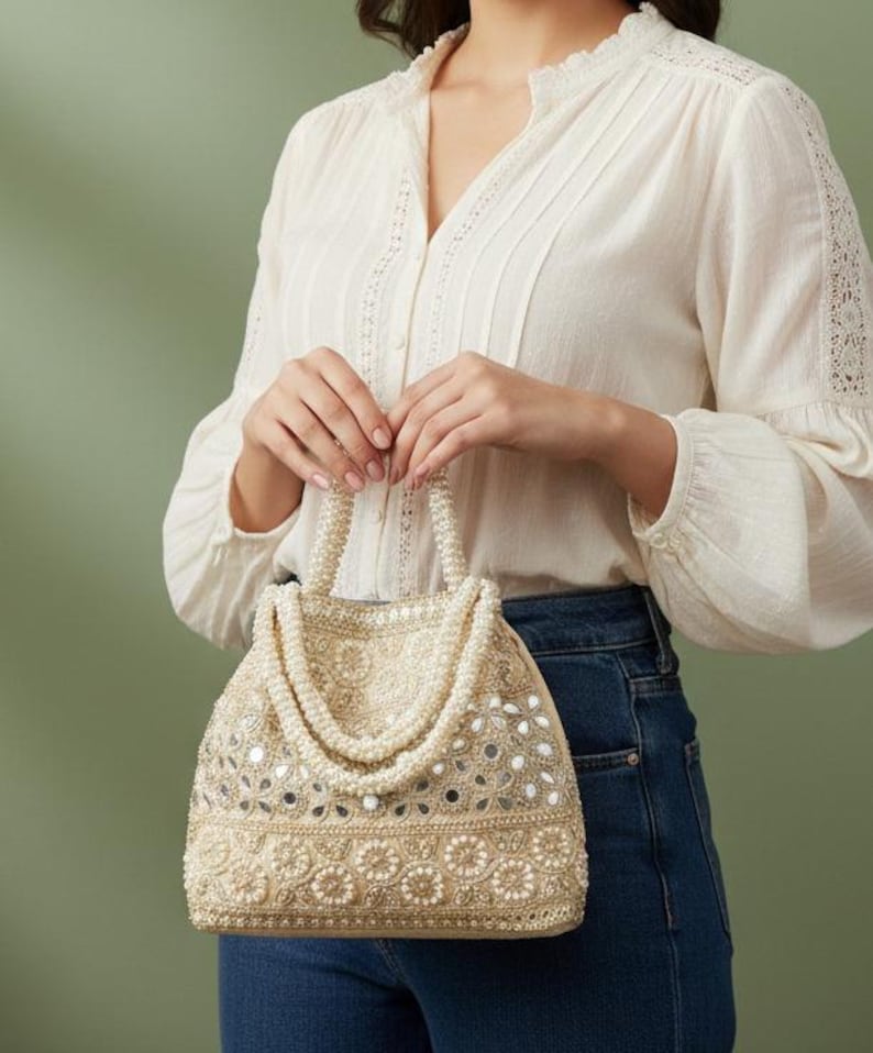 May include: A cream-coloured handbag with intricate beadwork and mirror accents. The bag features a pearl-beaded handle. The bag's design includes floral patterns and a structured shape. The person is wearing a white blouse and blue jeans.