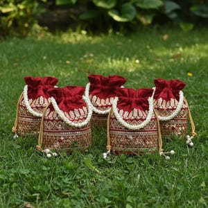 May include: Five maroon drawstring bags with gold and sequin embroidery. Each bag has a white beaded handle and a gold drawstring. The bags are arranged on green grass.